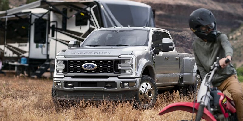 Gray Ford Super Duty truck parked in a field, an RV in the background, and a person on a dirt bike in the foreground
