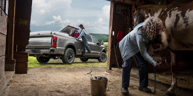 A woman works on a horse's hoof in a barn as another woman loads gear from a ford pickup truck outside